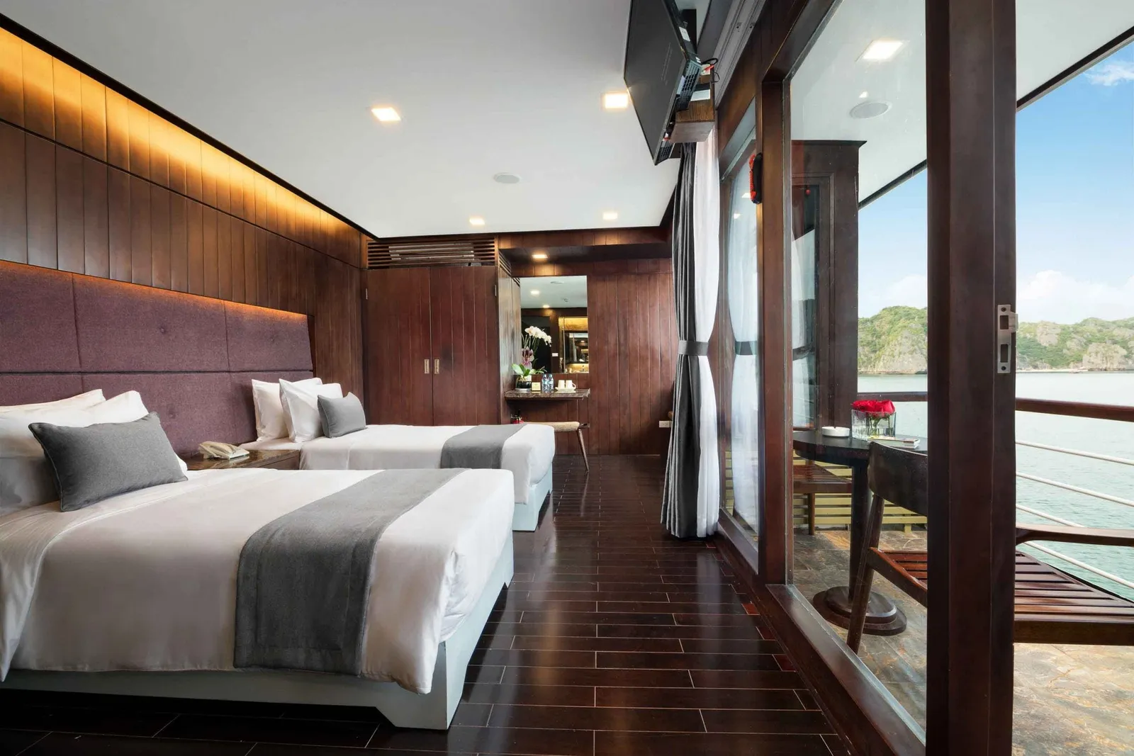 A cabin with two twin beds, dark wood paneling and floor, and open glass doors leading to a private balcony with a table and chairs overlooking karst limestone islands and calm water.