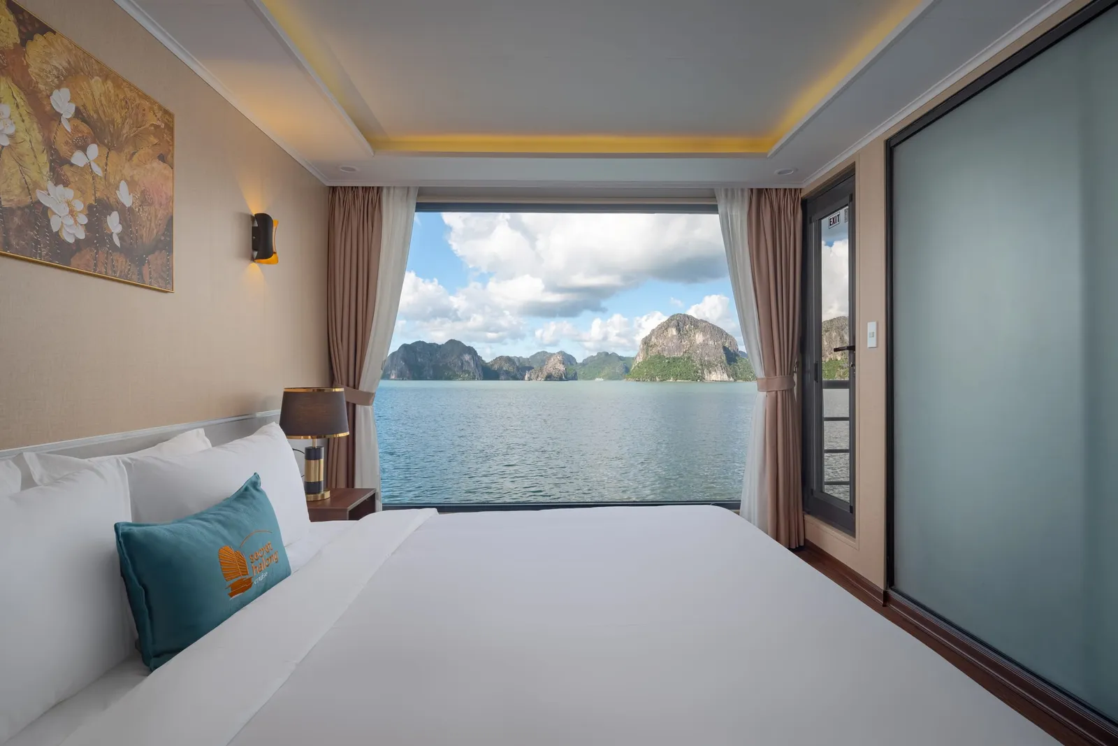 A double bed with a teal cushion facing a wide window with a view of the bay, with a tray ceiling and a framed painting.