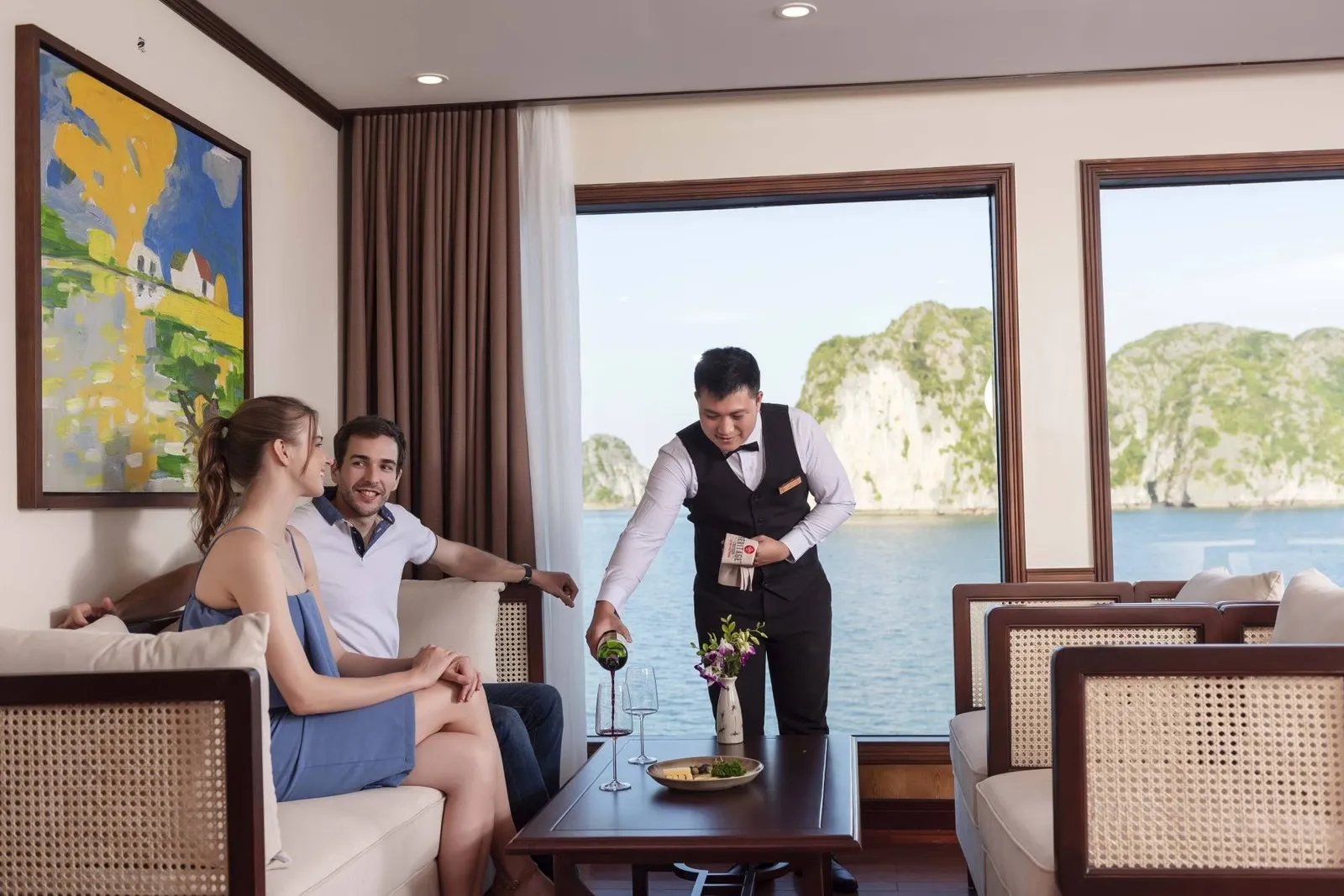 A staff member in vest and bow tie pours wine for two guests seated in a cabin lounge with large windows showing limestone karsts and bay water outside.