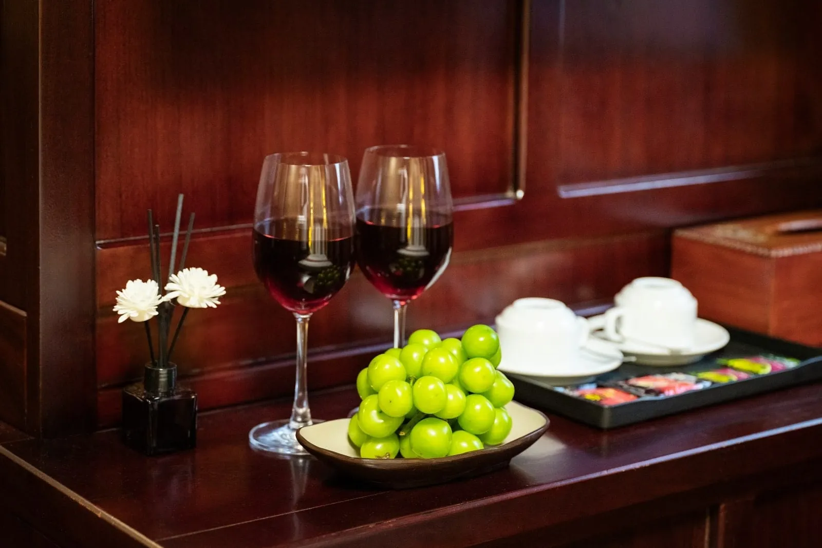 Two glasses of red wine, a dish of green grapes, a reed diffuser with white flowers, and a tray with cups and tea packets sit on a dark polished wooden cabinet against wood panelling.