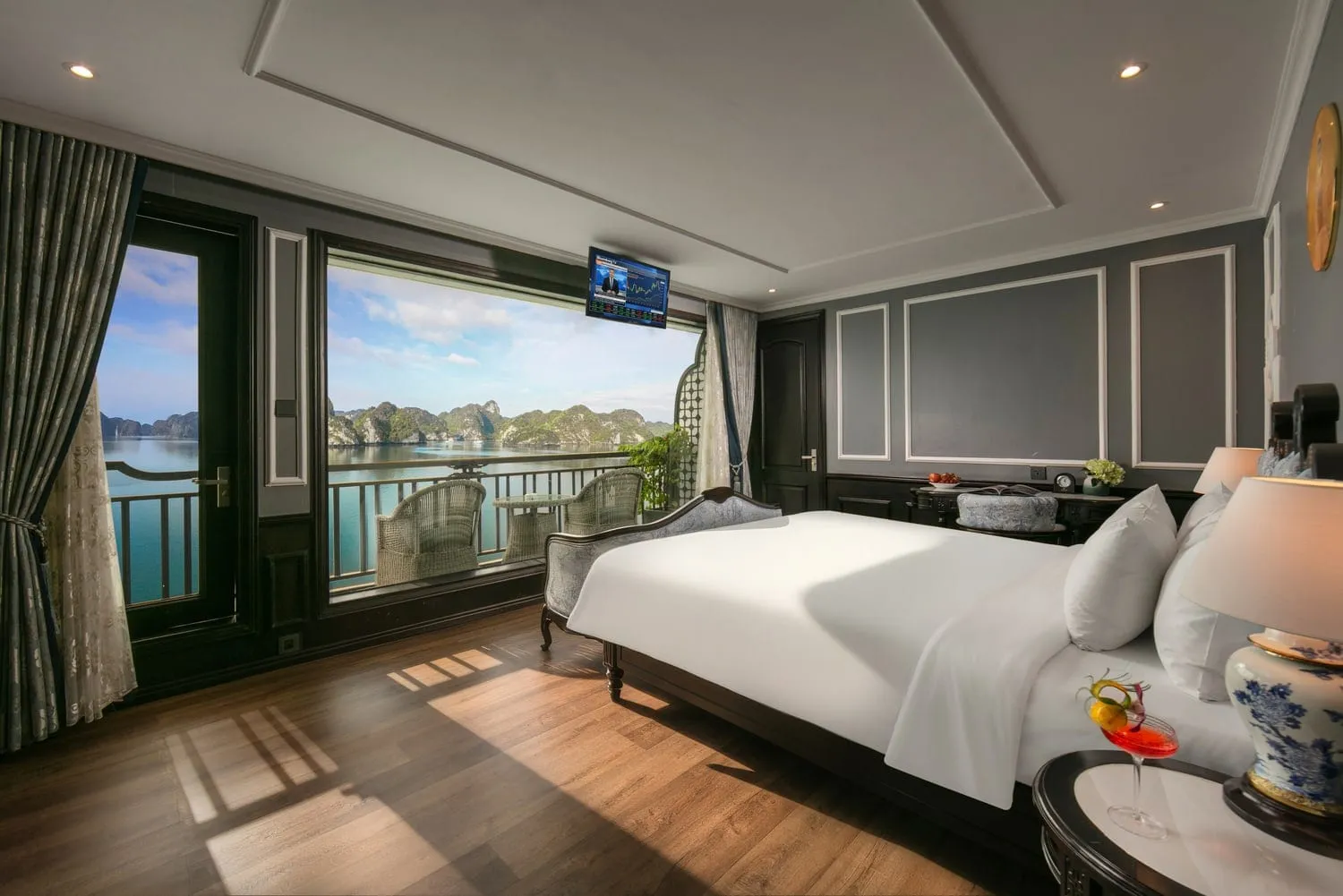 A luxury cabin interior with a white-sheeted bed, dark wood floors, and large windows revealing limestone karst islands and turquoise water outside.