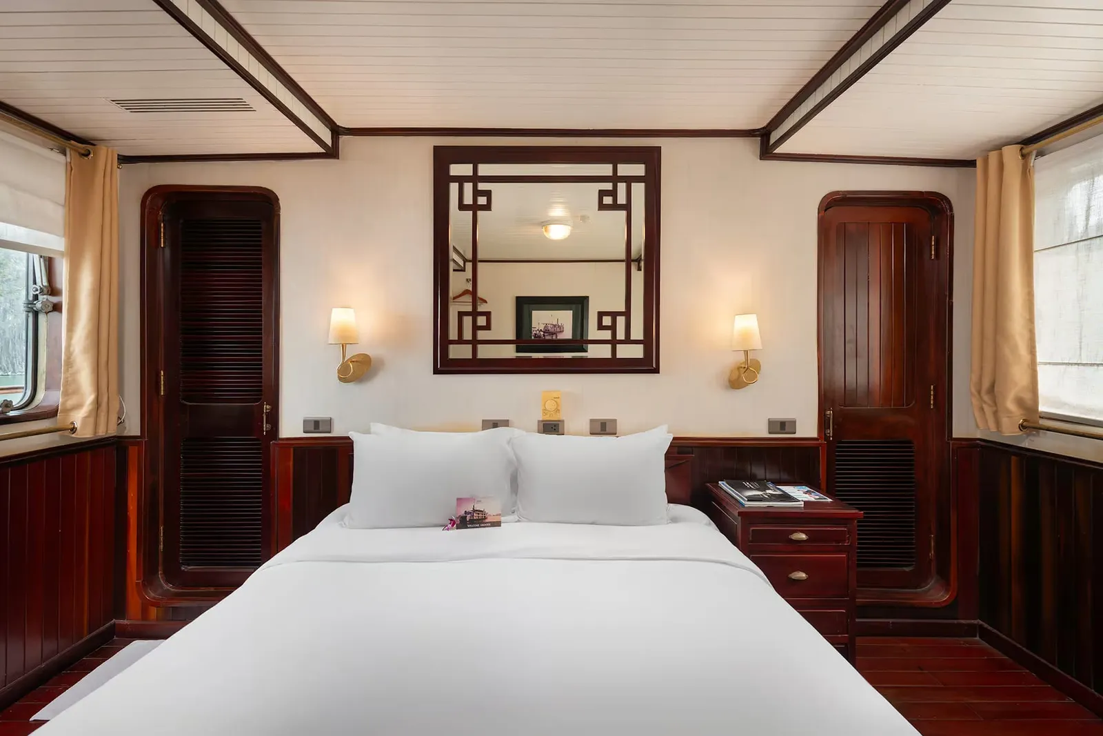 A cabin with a double bed in white linens, dark red-brown wood paneling and doors, a wall mirror with a lattice frame, two wall sconces, a bedside table with books, and windows with cream curtains showing greenery outside.