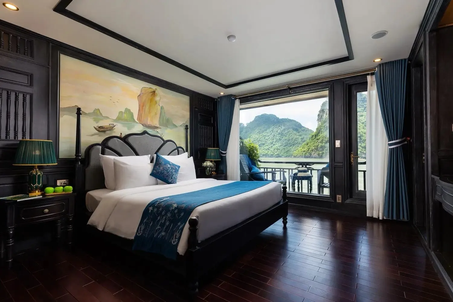 A dark-wood cabin with a king bed with teal runner, a painted mural of a sailing junk among karst islands, and a balcony door opening onto a view of limestone karst and calm water.