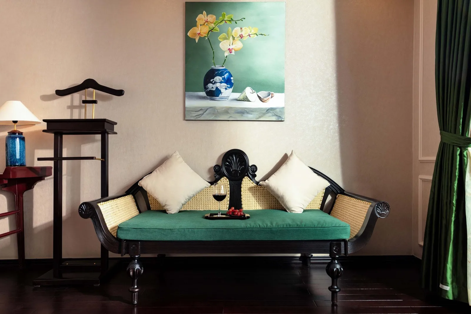 Interior room with a dark wood rattan daybed with green cushion and two cream pillows, a glass of red wine on a tray, an orchid painting on the wall, a valet stand, a side table with lamp, and green silk curtains.