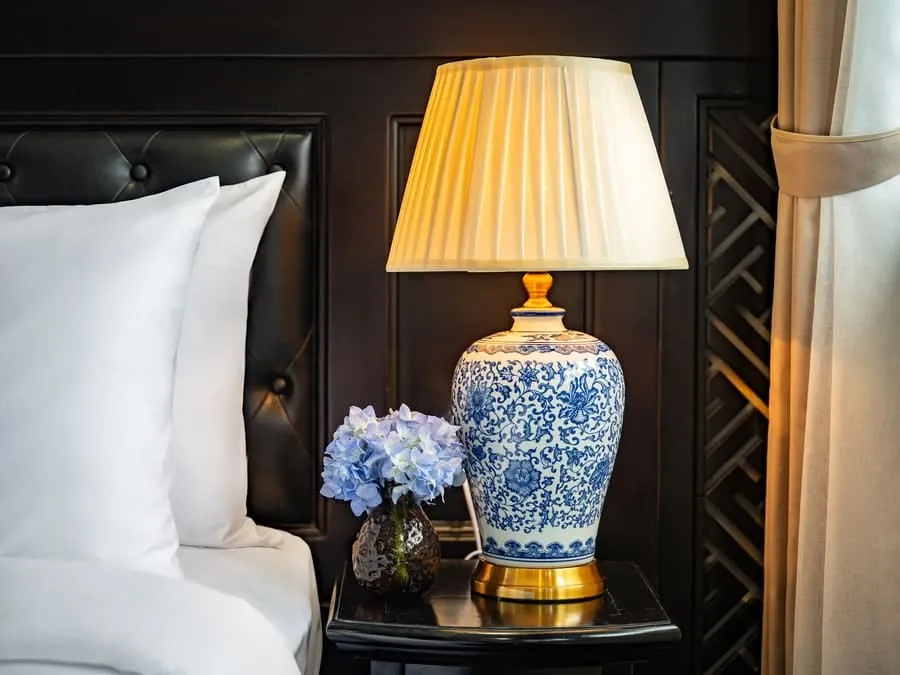 A blue-and-white porcelain lamp and flowers on a bedside table next to white pillows.