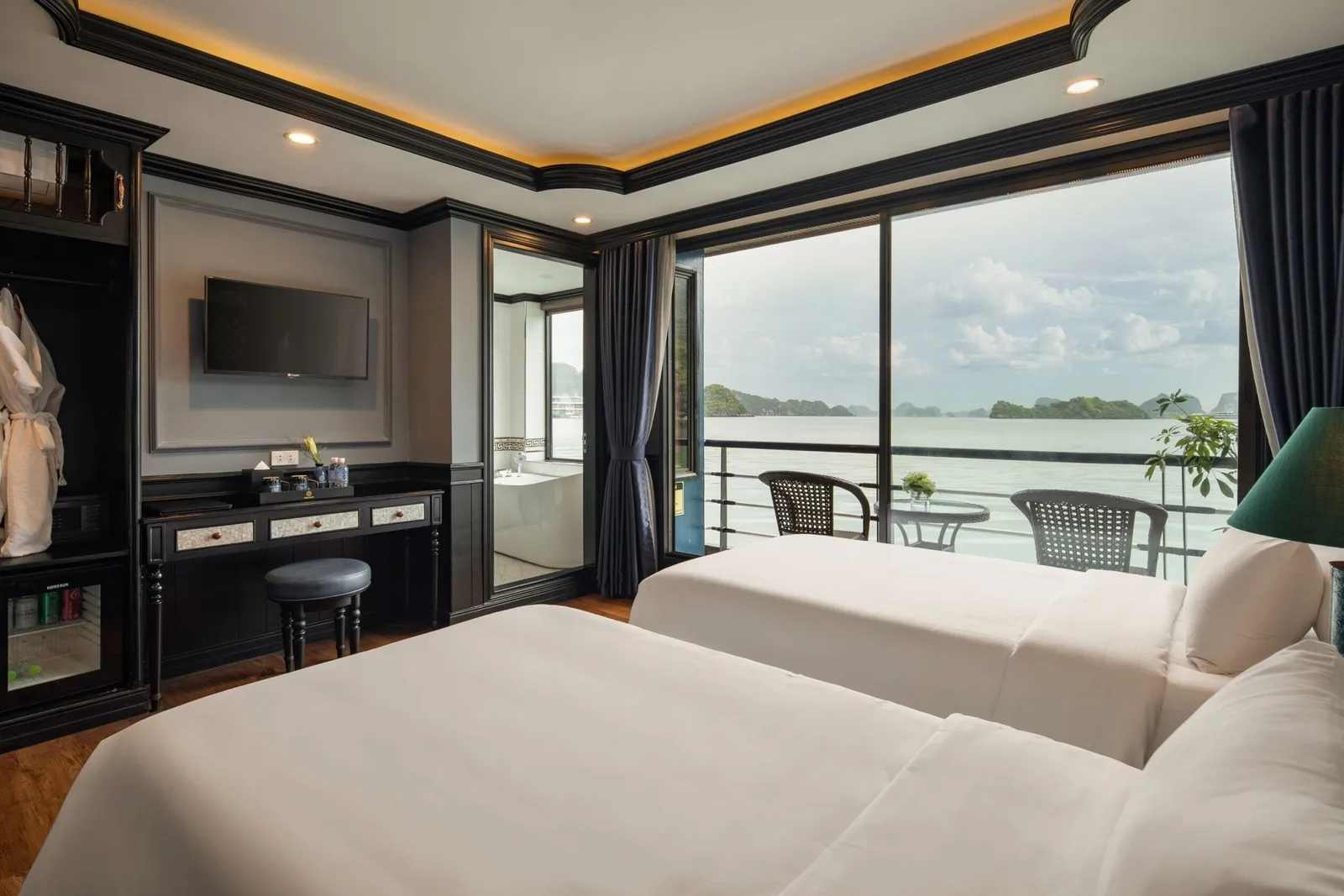 A luxury cabin interior with two white beds, dark wood furniture, and large windows opening to a balcony overlooking calm water with limestone karst islands in the distance.