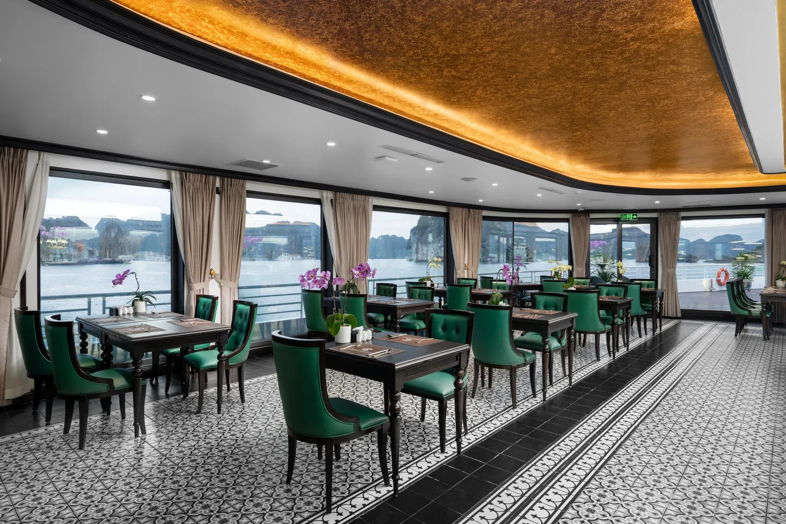 A dining room interior with green velvet chairs, patterned tile floor, curved gold ceiling, and panoramic windows showing limestone karsts and water outside.