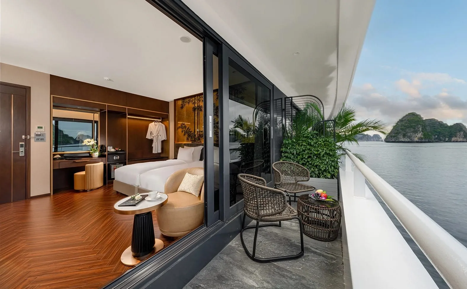 A cruise cabin with twin beds, herringbone wood floor and a bathrobe, opening through sliding glass doors onto a private balcony with wicker chairs, plants and a fruit bowl, overlooking calm water and a limestone karst island.