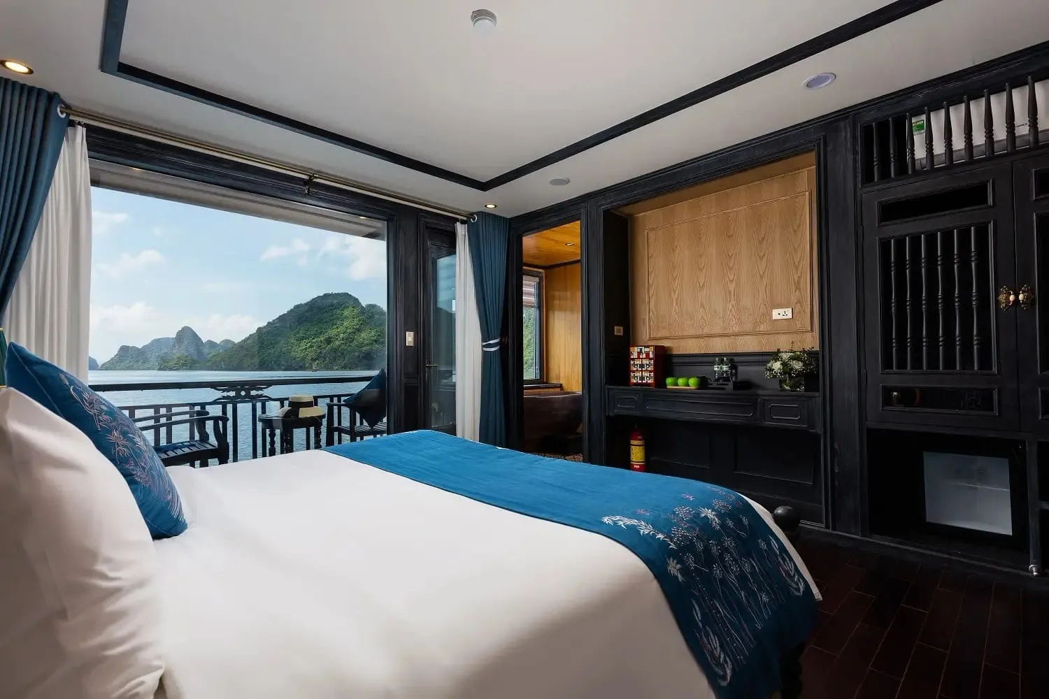 A cabin with a white bed, blue embroidered runner and pillows, dark wood cabinetry, and a balcony with chairs overlooking a bay with green karst islands.