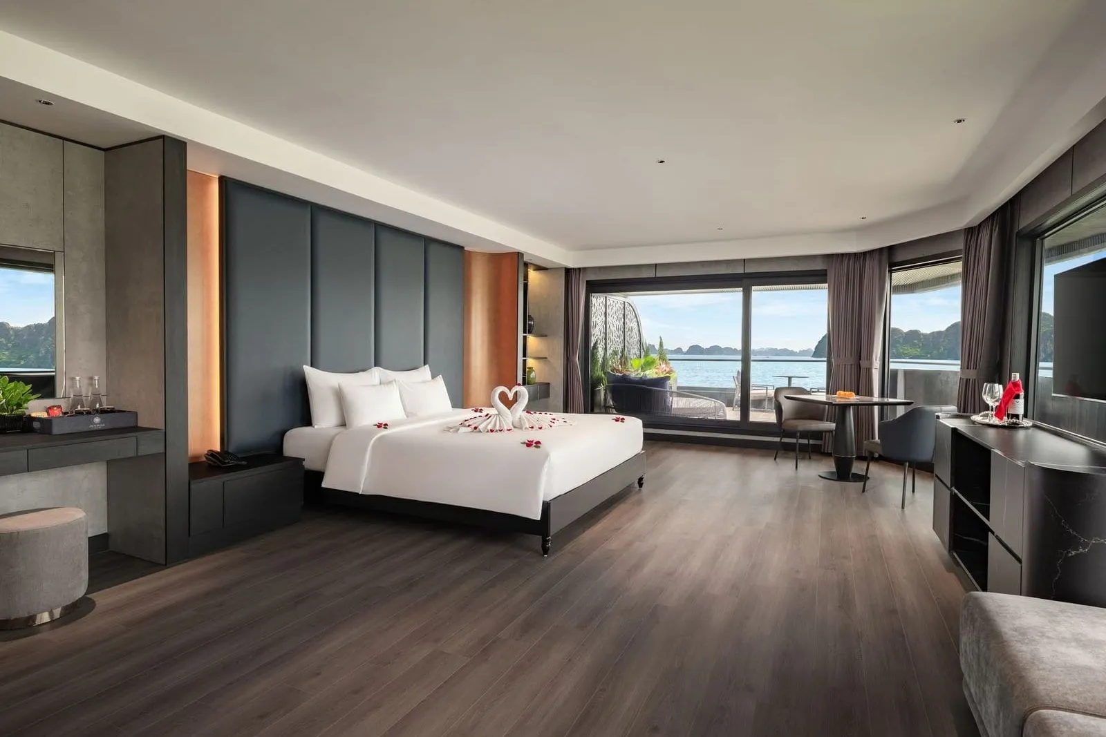 A spacious cabin with a large bed decorated with towel swans and rose petals, a dining table and sliding glass doors opening onto a balcony with a view of calm water and limestone karst islands.