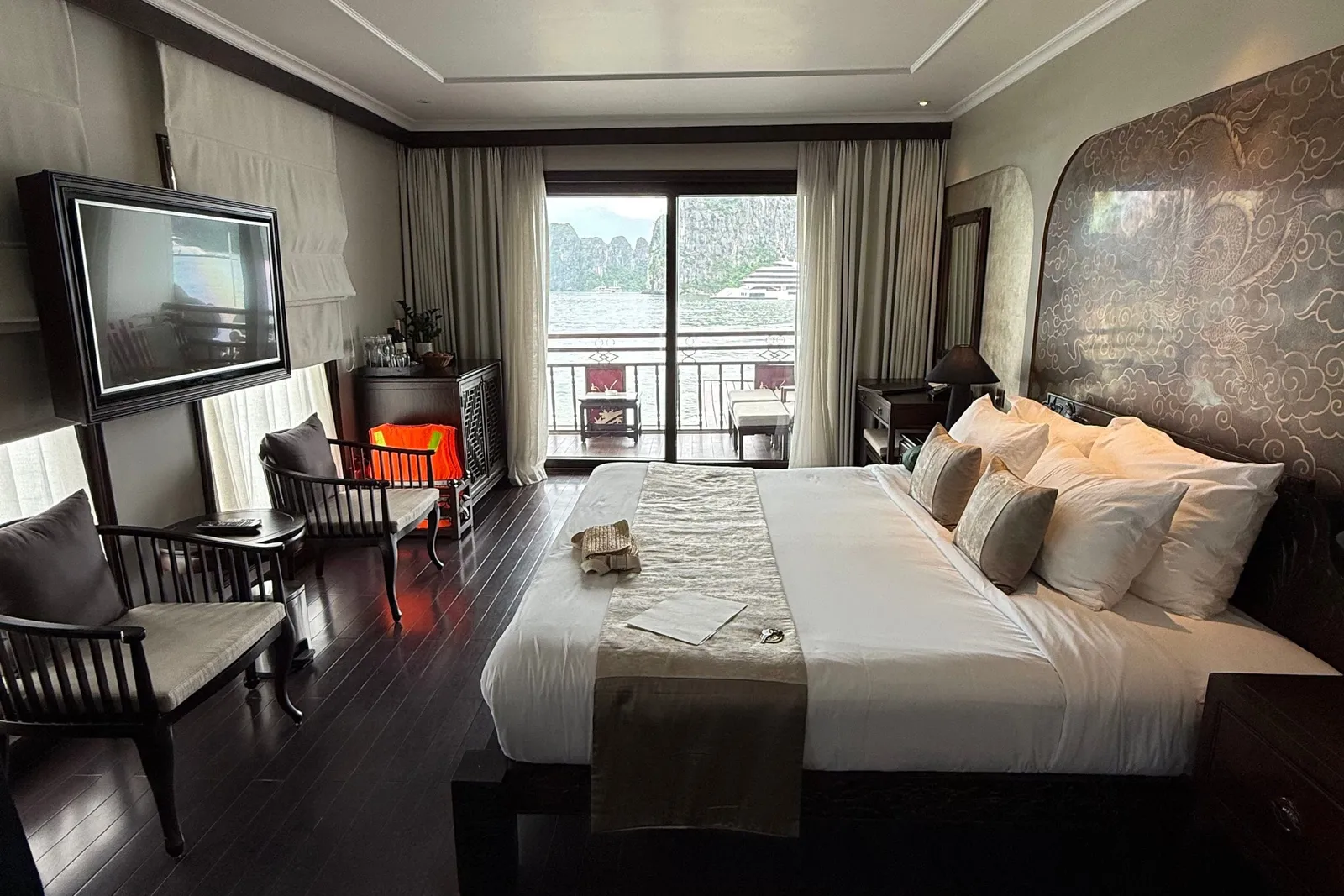 A dark-wood cruise cabin with a king bed, a carved dragon headboard, armchairs, a wall-mounted TV and an orange life jacket, with a balcony door opening onto karst limestone cliffs and water.