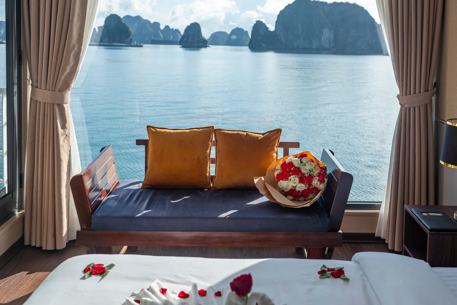 A cabin window seat with orange cushions beside a large window with curtains and a bay view, with the foot of the bed in front.
