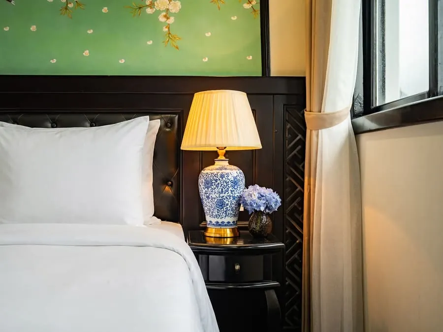 A bed corner with white pillows, a porcelain lamp and a curtain beside a window.