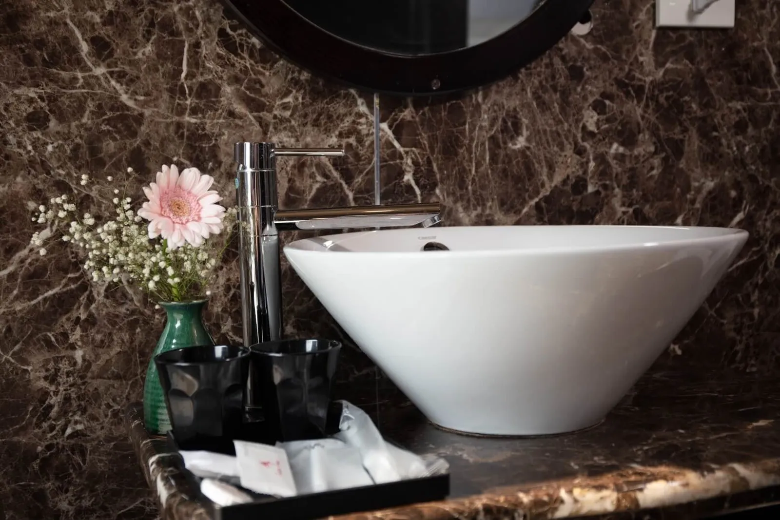 A white vessel sink with a chrome tap on a dark brown marble counter, with a small green vase holding a pink gerbera and baby's breath, two black cups and a tray of wrapped amenities, beneath a round dark-framed mirror.