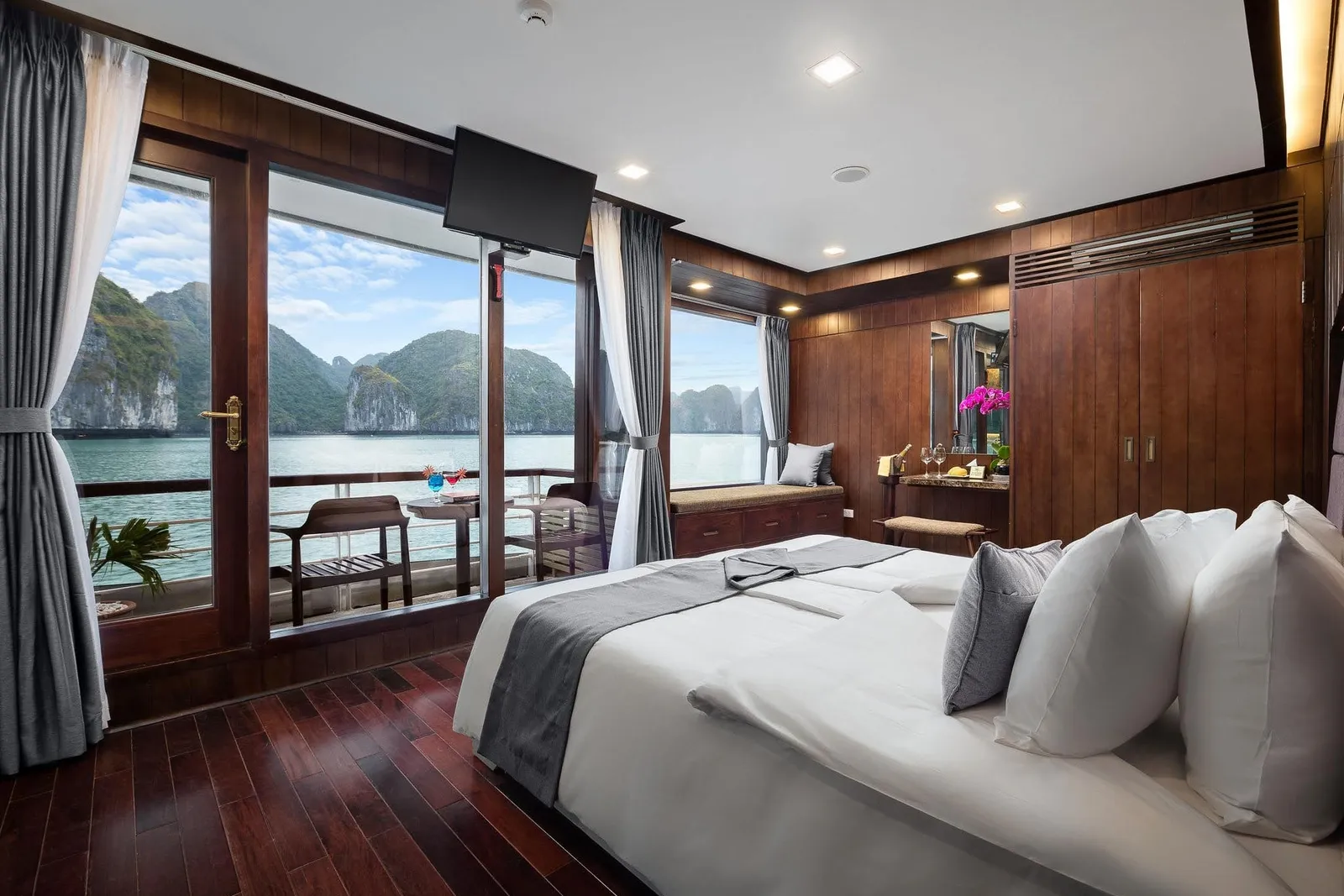 A wood-paneled cabin with a double bed, a TV, and glass doors opening to a private balcony overlooking limestone karst islands on calm water.