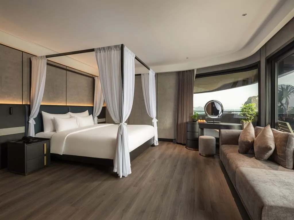 A modern cabin with a canopy bed with white drapes, a grey sofa, a vanity with round mirror and a curved panoramic window showing limestone karst peaks outside.