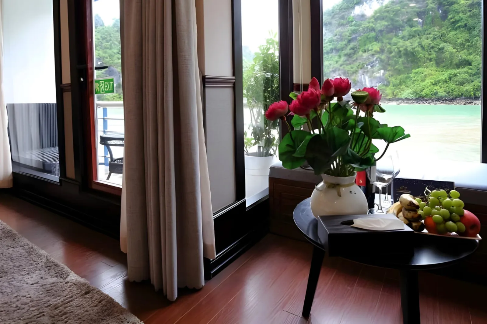 A cabin corner with a round table, roses and fruit by a window and balcony doors.