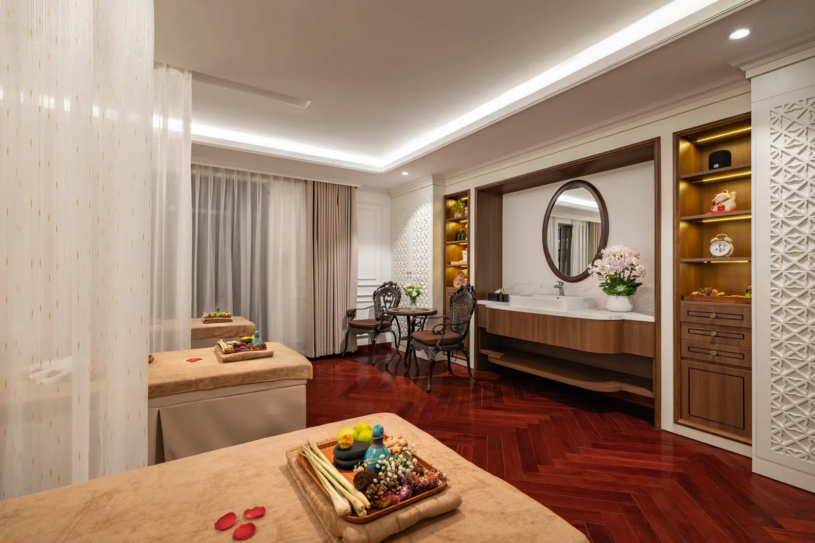 A luxurious spa or massage cabin interior with two massage tables, herringbone wood floors, vanity area with mirror, and ambient lighting.