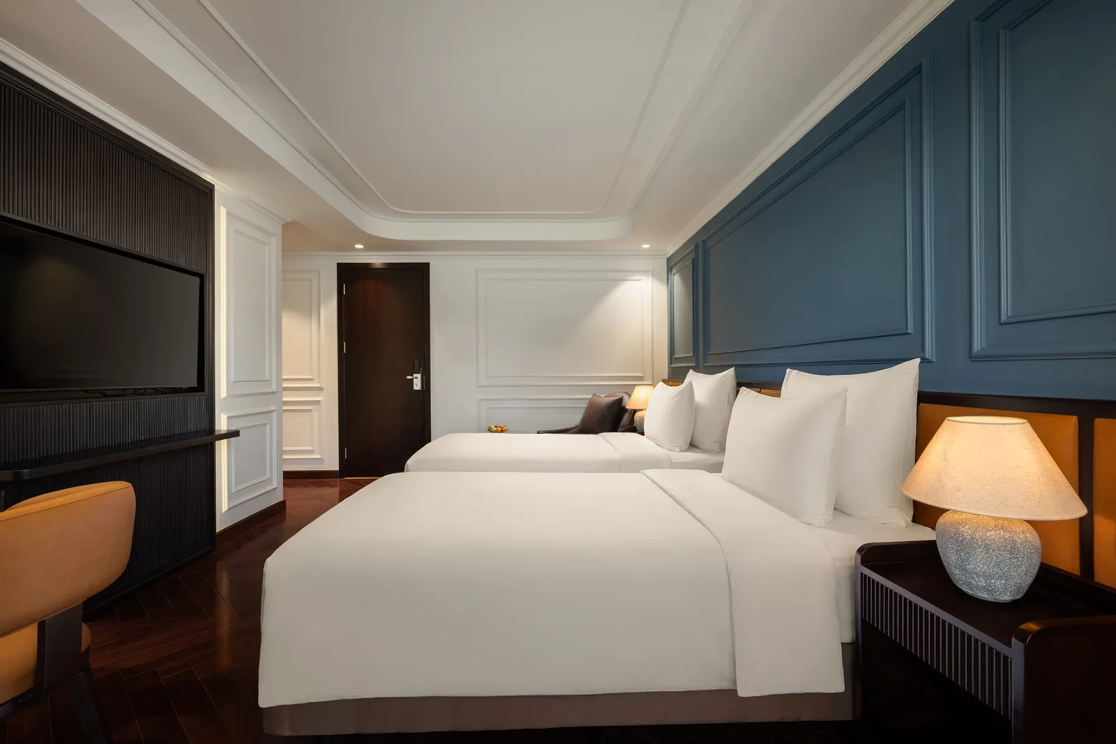 A cabin with two white-linen twin beds, a wall-mounted TV, blue and white paneled walls, dark herringbone wood floor, a table lamp and an orange chair.