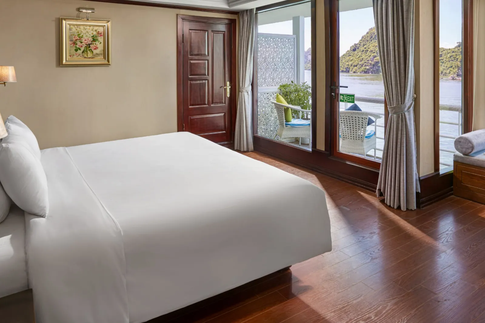 A large bed with white linen next to balcony doors and a curtain, with a wooden door at the back.