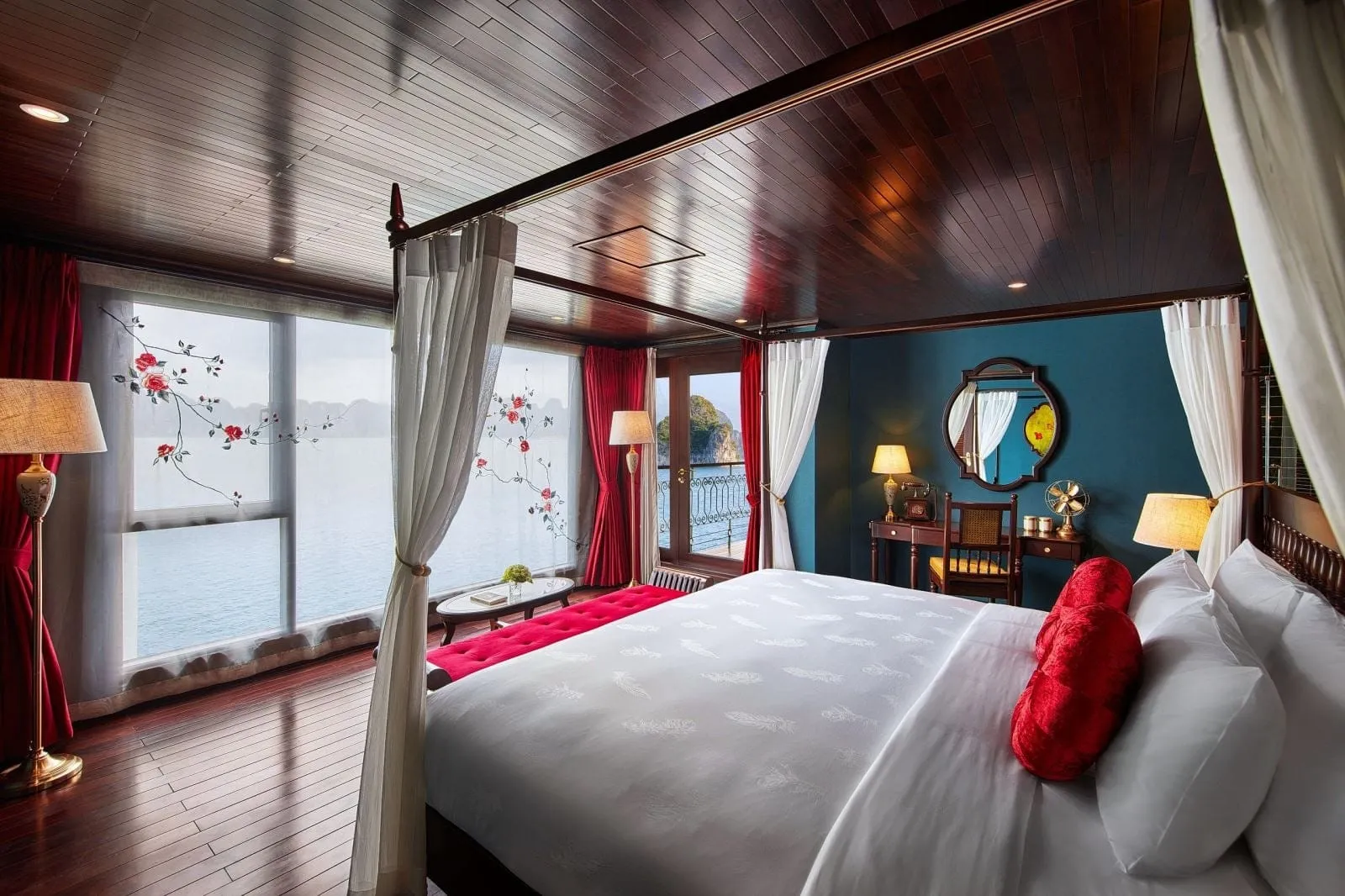 A cabin with a four-poster bed, dark wood ceiling and floor, red curtains and cushions, and large windows and a balcony door looking out on calm water and limestone karst islands.