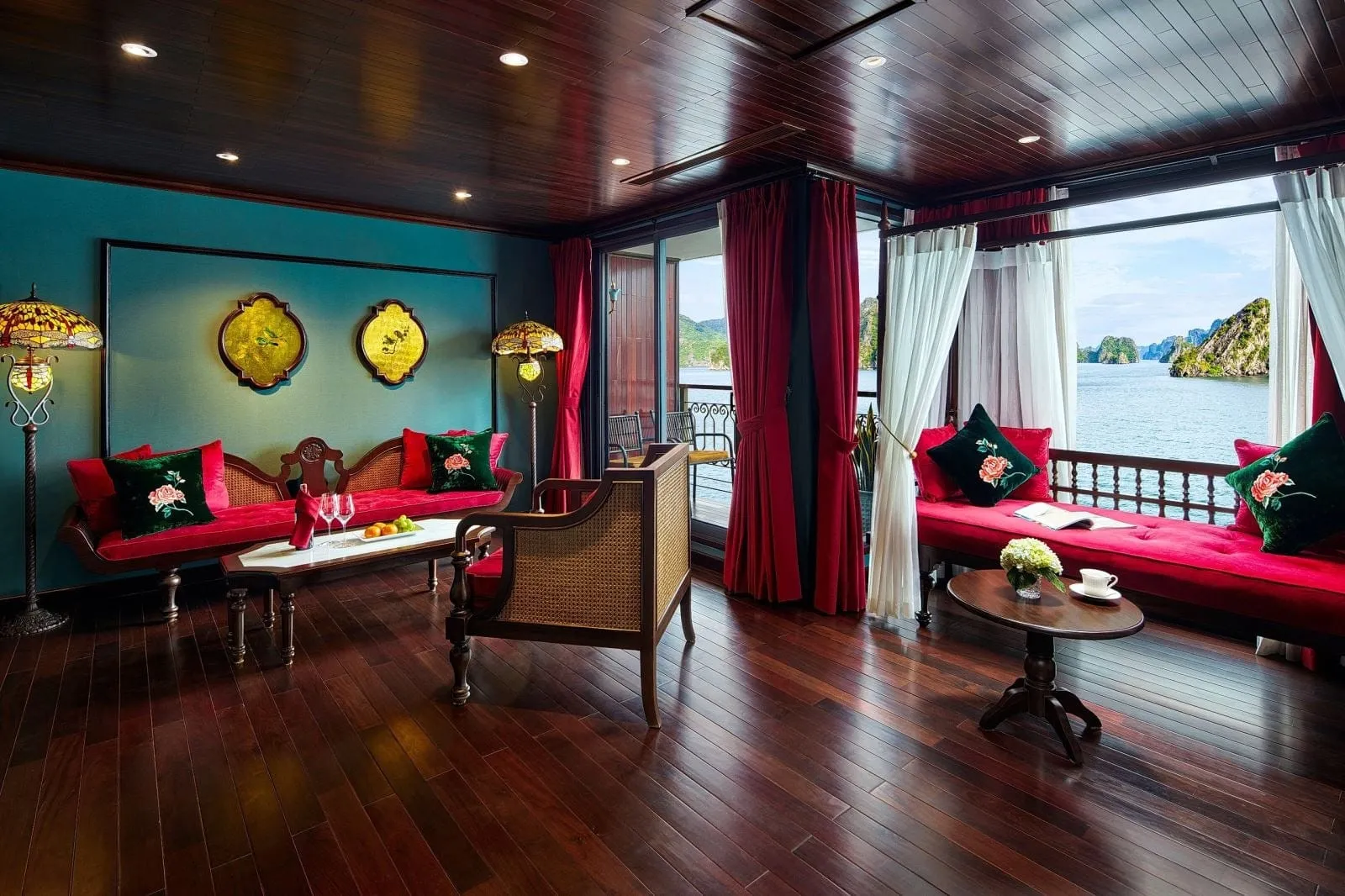 A wood-paneled suite or lounge with red velvet sofas, green embroidered cushions, Tiffany-style floor lamps and red curtains, with large windows and a balcony looking out to limestone karsts on calm water.