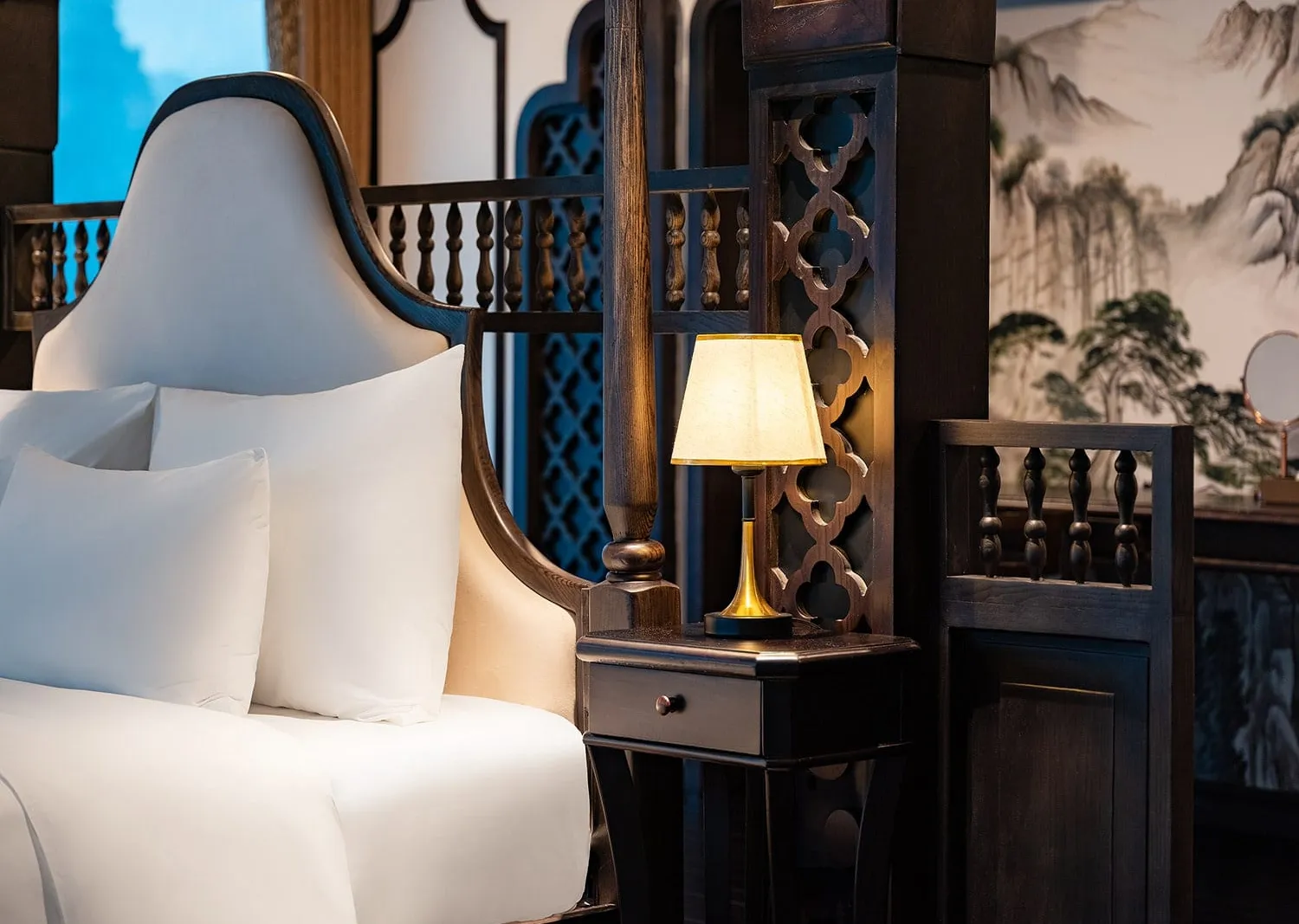 Close-up of a cabin bed with white pillows and a dark wood four-poster frame, a lit bedside lamp on a small nightstand, carved lattice screens, and an ink landscape mural on the wall.