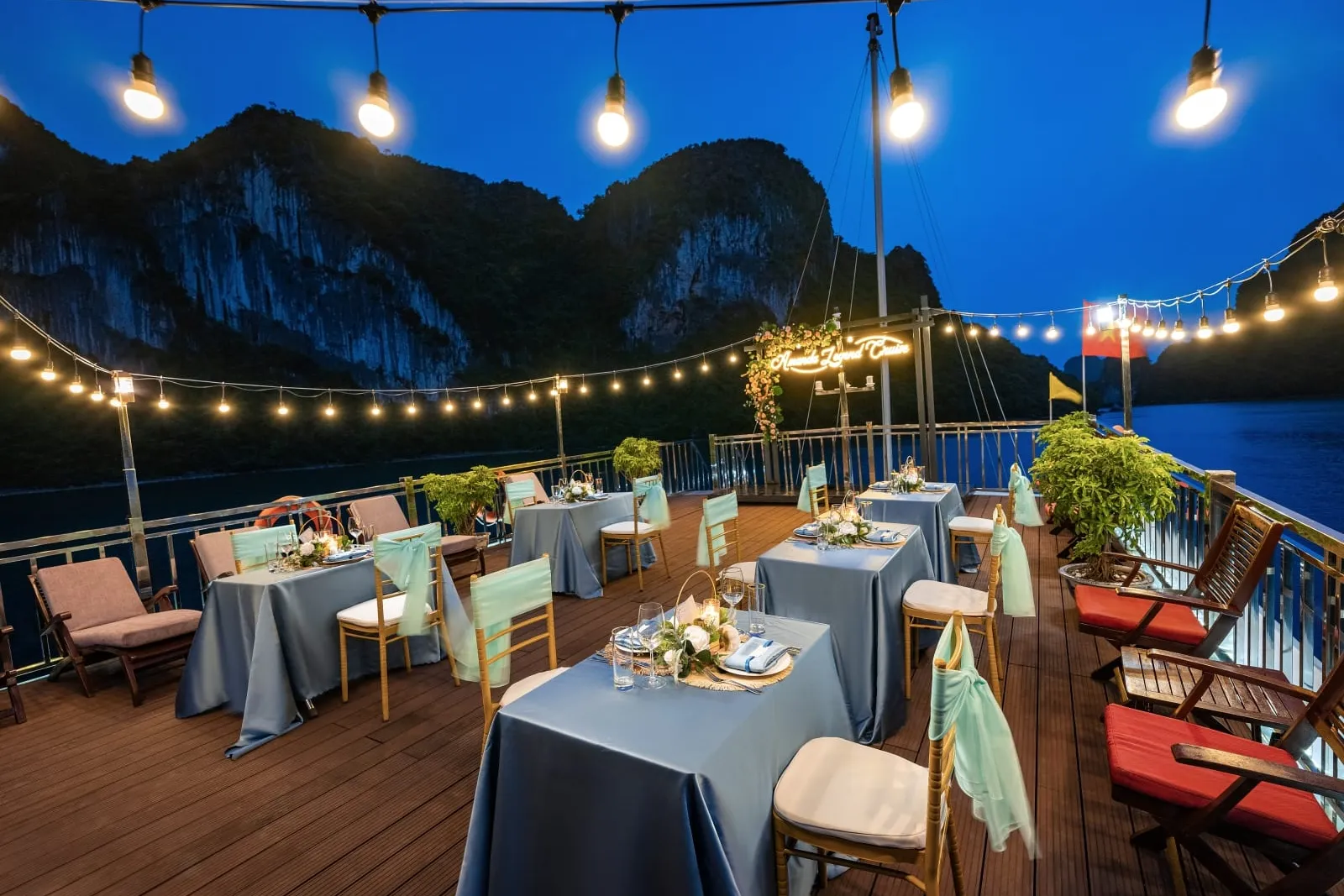 A dusk view of a boat's upper deck set with square tables in grey-blue linens and teal-sashed chairs, under string lights with a lit neon sign on a floral arch, with karst cliffs and dark water behind.