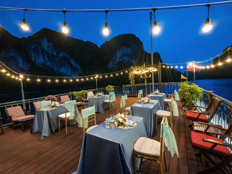 A dusk view of a boat's upper deck set with square tables in grey-blue linens and teal-sashed chairs, under string lights with a lit neon sign on a floral arch, with karst cliffs and dark water behind.