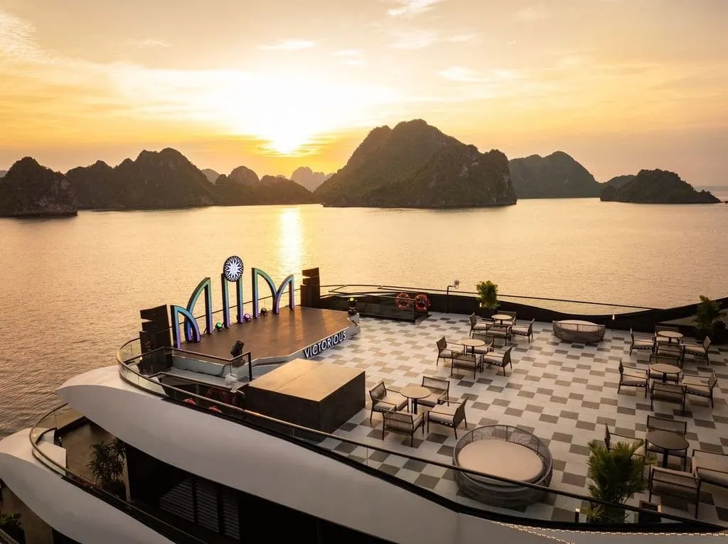 A sunset view from an upper level of a cruise vessel over a checkerboard-tiled sun deck with tables, chairs and a stage bearing the word 'VICTORIOUS' and a sign-like arched logo, with limestone karst islands across the water.