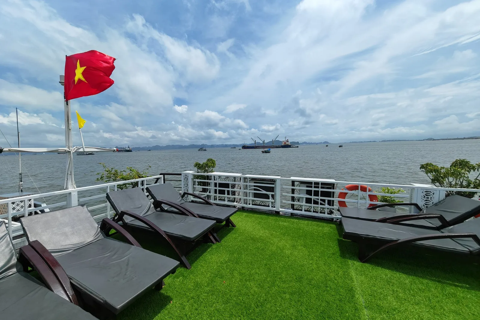 A sun deck with lounge chairs on artificial grass, Vietnamese flag flying, overlooking calm waters with distant limestone karsts and vessels.