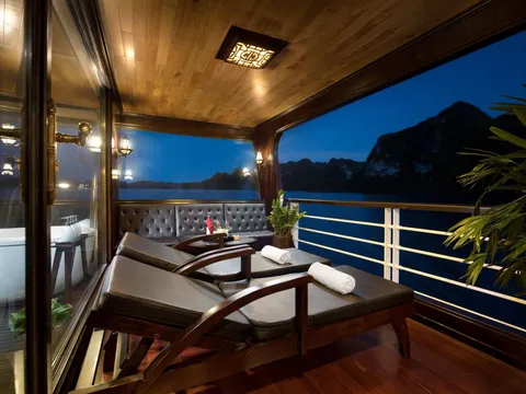 A covered private balcony on a boat at dusk with wooden loungers, rolled white towels, a tufted bench, a bathtub behind glass, and limestone karst silhouettes across the water.