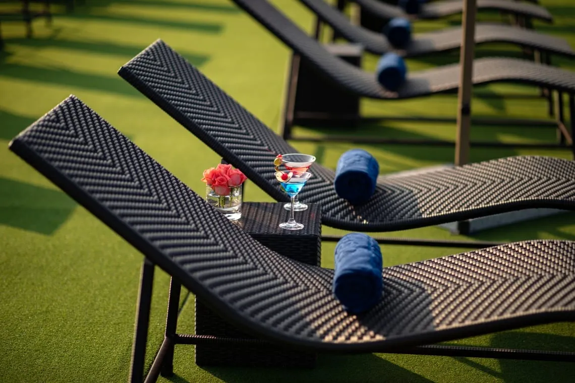 A row of dark woven loungers with rolled blue towels on artificial grass, with a small side table holding two cocktails and a glass of pink roses.