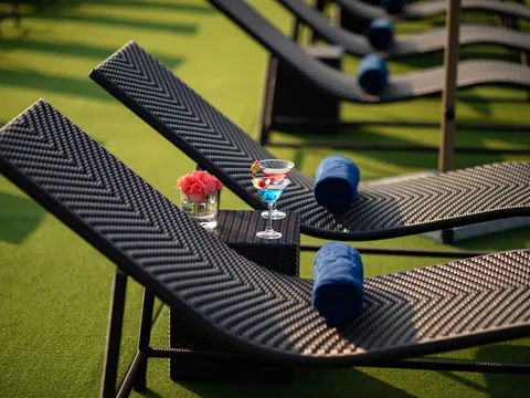 A row of dark woven loungers with rolled blue towels on artificial grass, with a small side table holding two cocktails and a glass of pink roses.