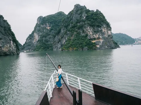A person stands on a boat's bow deck with white railings facing limestone karsts rising from calm green water.