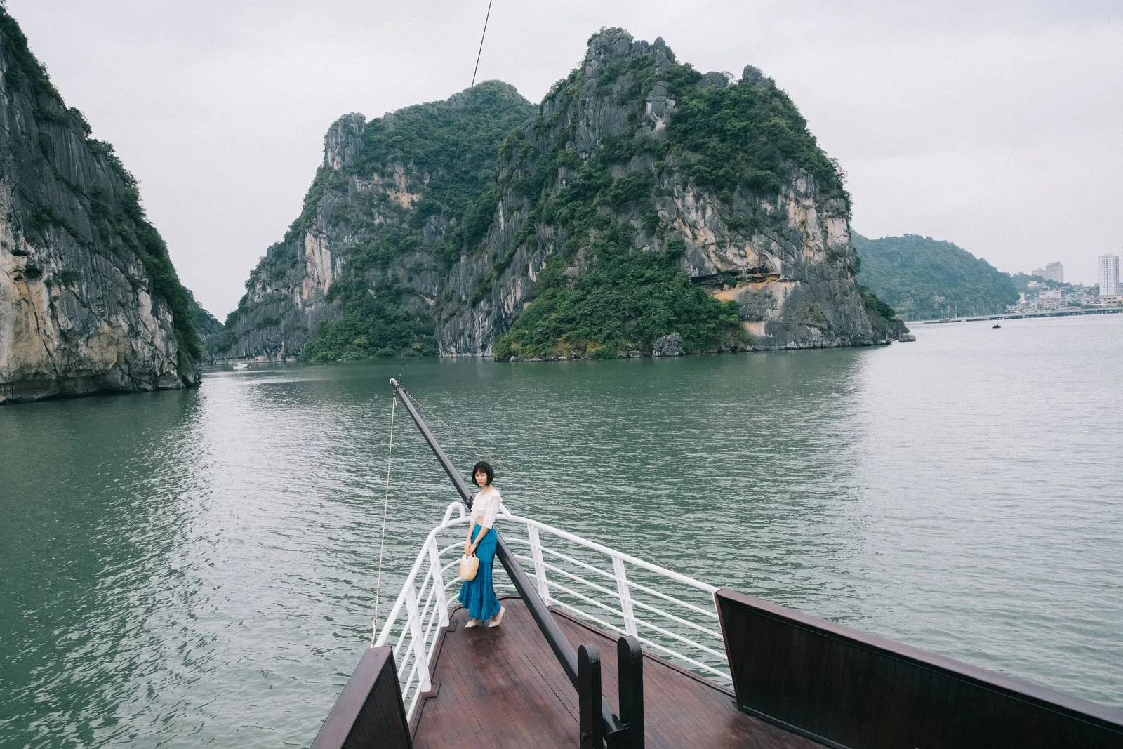 A person stands on a boat's bow deck with white railings facing limestone karsts rising from calm green water.