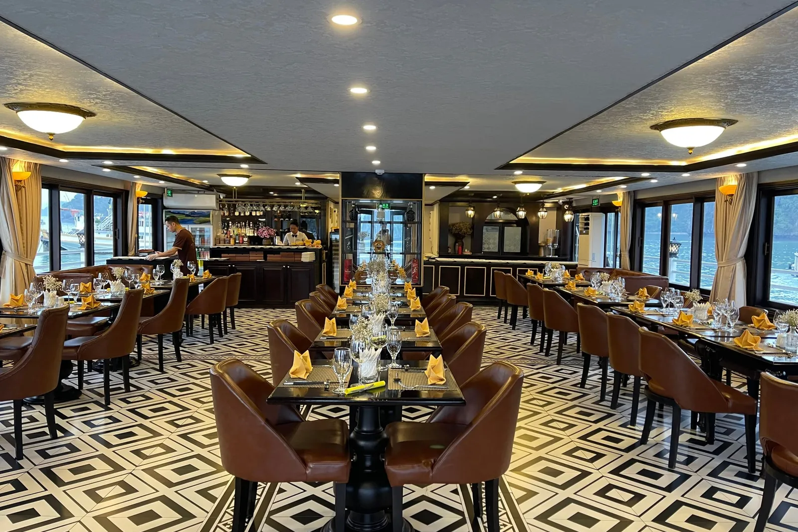 Interior of a ship's dining room with set tables, orange napkins, brown leather chairs, a geometric black-and-white floor, a bar with a staff member at the back, and windows showing water and boats outside.