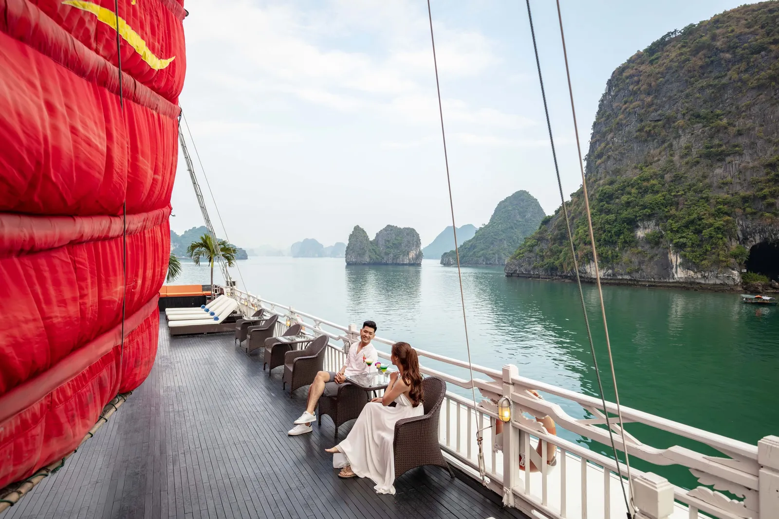 A man and a woman sit at a small table with drinks on the wooden deck of a junk boat with a large red sail, beside a white railing, overlooking green water and limestone karst islands.