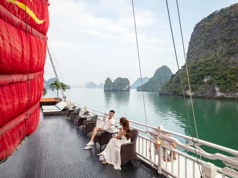 A man and a woman sit at a small table with drinks on the wooden deck of a junk boat with a large red sail, beside a white railing, overlooking green water and limestone karst islands.