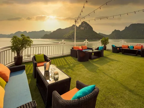 A sundeck with artificial grass and dark wicker sofas, armchairs and tables with colorful cushions, string lights overhead and a white railing, facing limestone karst islands on calm water at sunset.