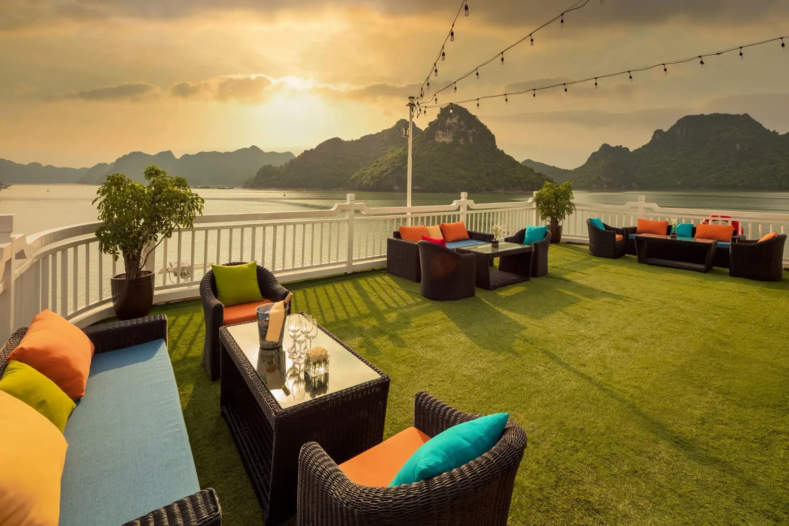 A sundeck with artificial grass and dark wicker sofas, armchairs and tables with colorful cushions, string lights overhead and a white railing, facing limestone karst islands on calm water at sunset.