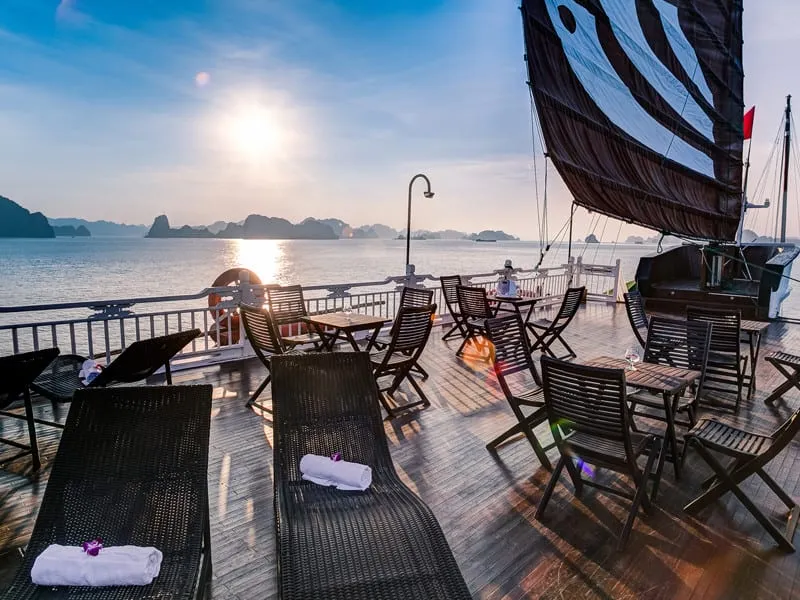 A sun deck with loungers, cafe chairs and tables and a dark sail, with the sun over the bay.