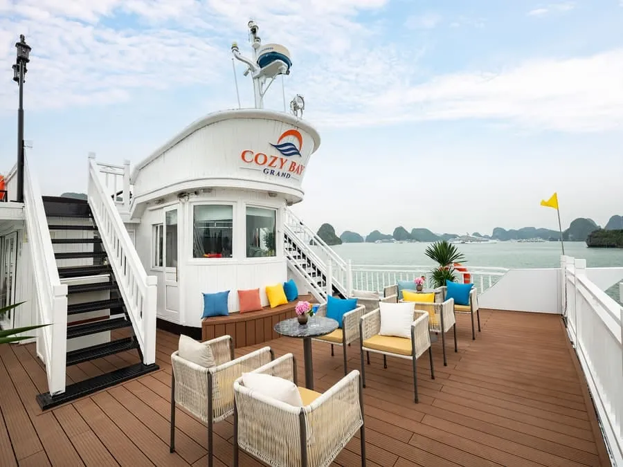 The upper deck with the white wheelhouse lettered Cozy Bay Grand, wicker armchairs and cushions on a timber deck.