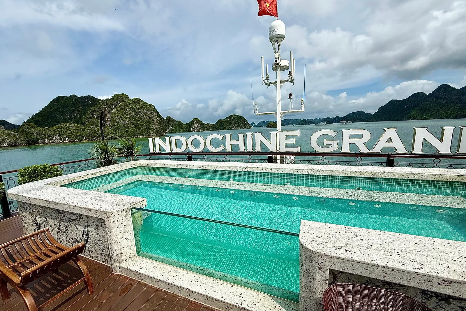 A swimming pool on a boat deck with large three-dimensional letters spelling INDOCHINE GRAN mounted on the railing, with limestone karsts and water in the background.
