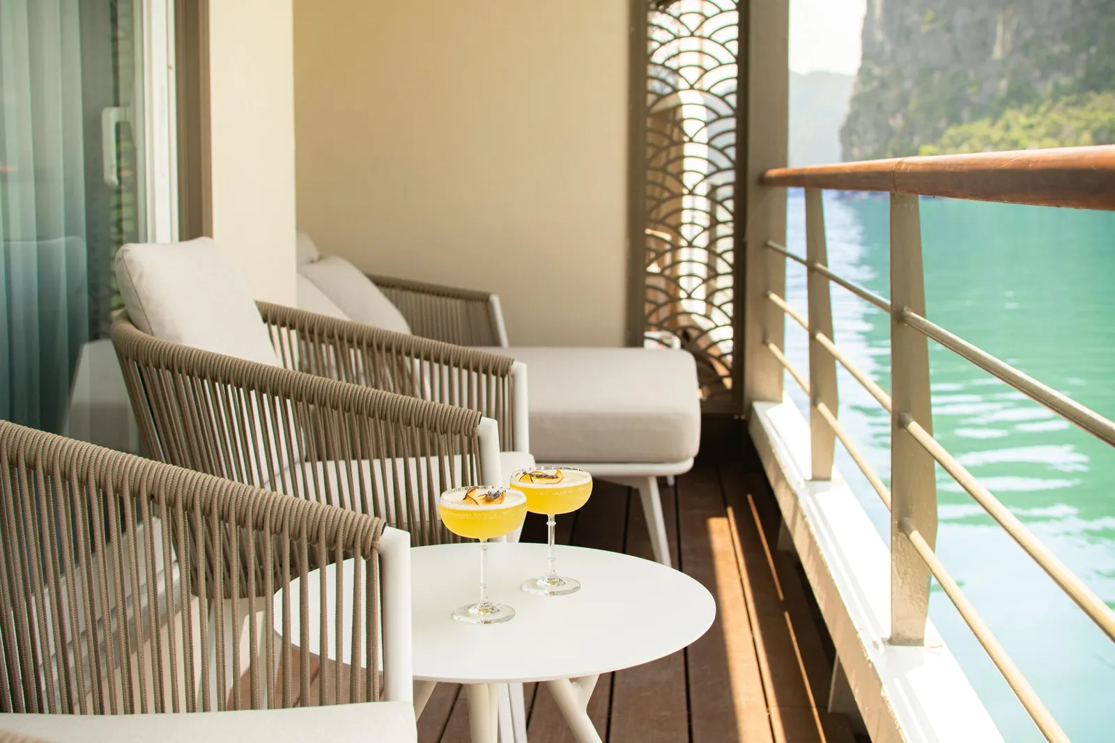 A private balcony or deck with wicker furniture, two yellow cocktails on a white table, wooden railing, and turquoise water with limestone karsts visible in the background.