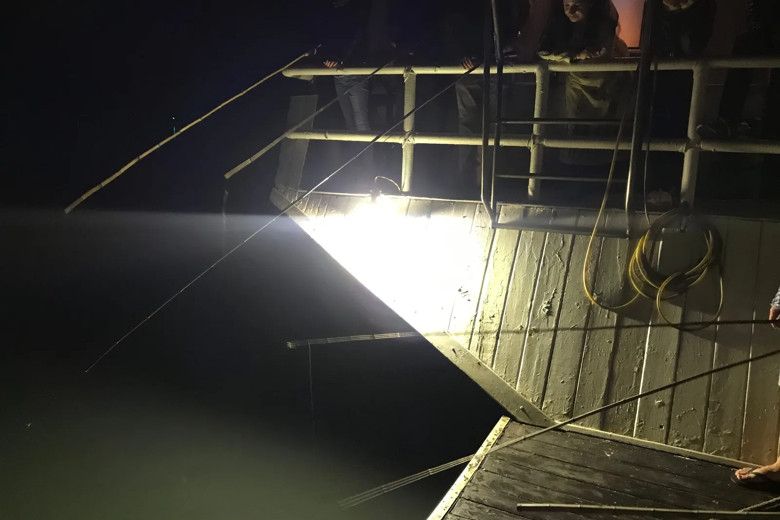 A wooden boat deck at night illuminated by a single bright spotlight shining on the water with a person visible at the railing.