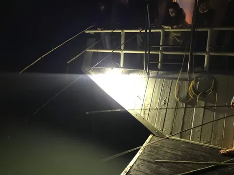 A wooden boat deck at night illuminated by a single bright spotlight shining on the water with a person visible at the railing.