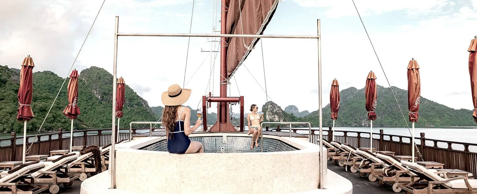 Two women relax around a small tiled pool on the sun deck of a wooden boat, with closed red parasols, loungers, a red sail, and green limestone karst islands across the water behind them.