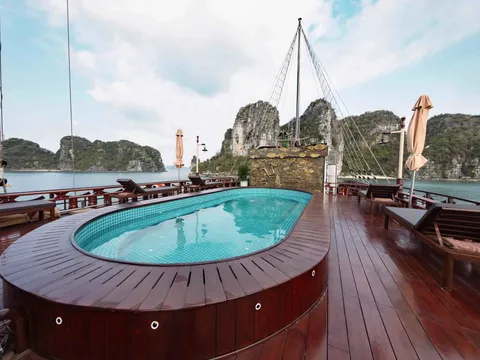 A wooden sun deck with an oval tiled plunge pool, loungers and closed umbrellas, with rigging and a gilded relief panel, and green limestone karst islands across the water under a partly cloudy sky.