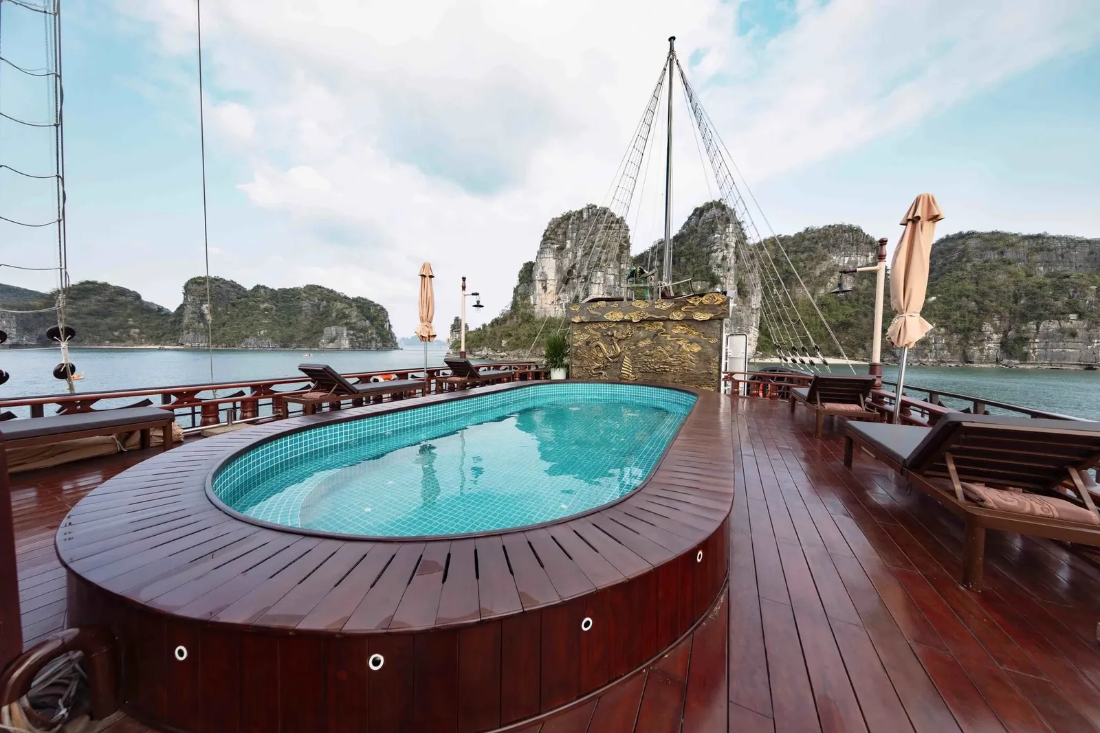 A wooden sun deck with an oval tiled plunge pool, loungers and closed umbrellas, with rigging and a gilded relief panel, and green limestone karst islands across the water under a partly cloudy sky.
